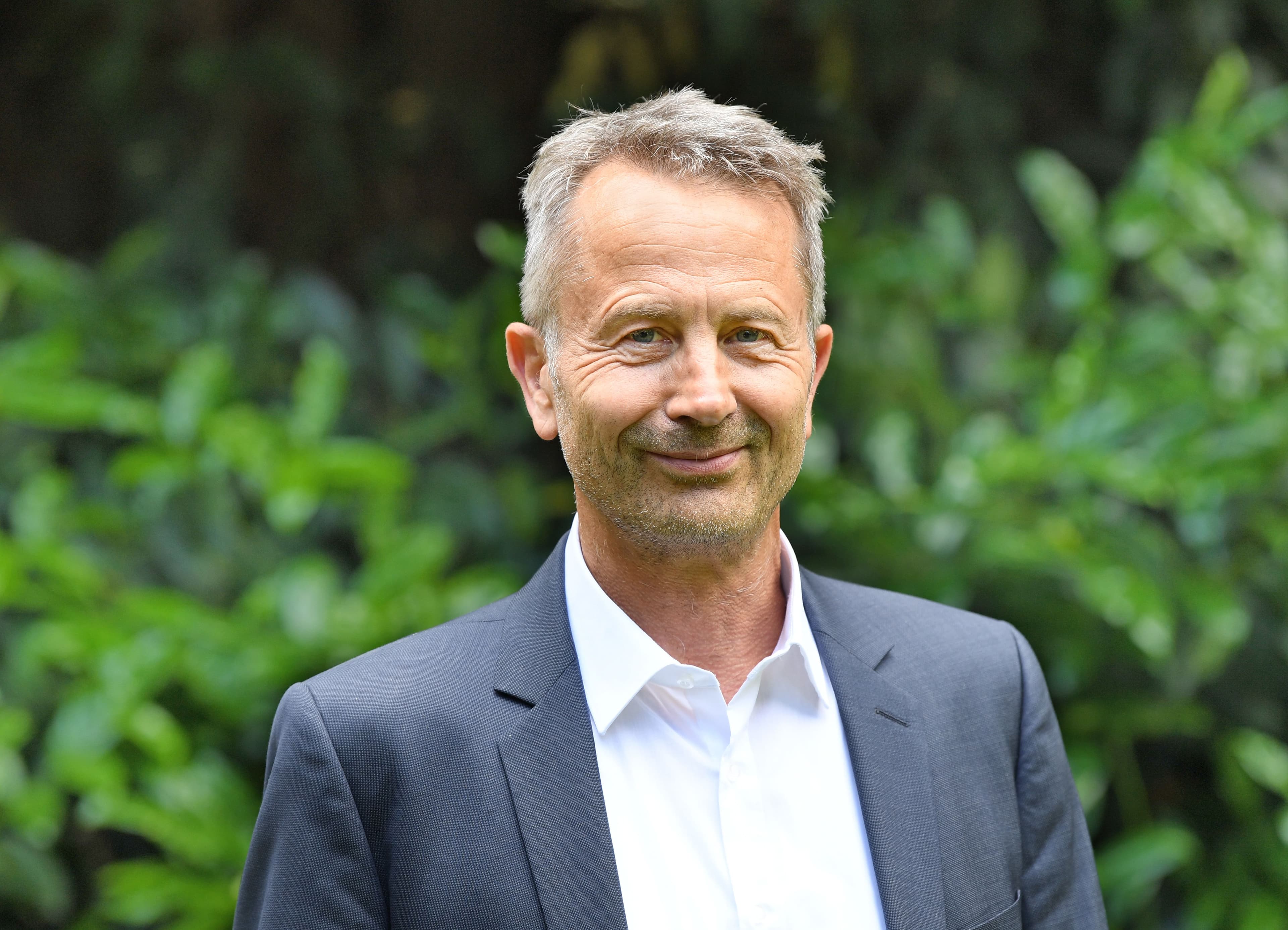 Porträt von Thomas Hertfelder in einem dunklen Anzug mit weißem Hemd. Er steht im Freien vor einem unscharfen Hintergrund aus grünen Bäumen.