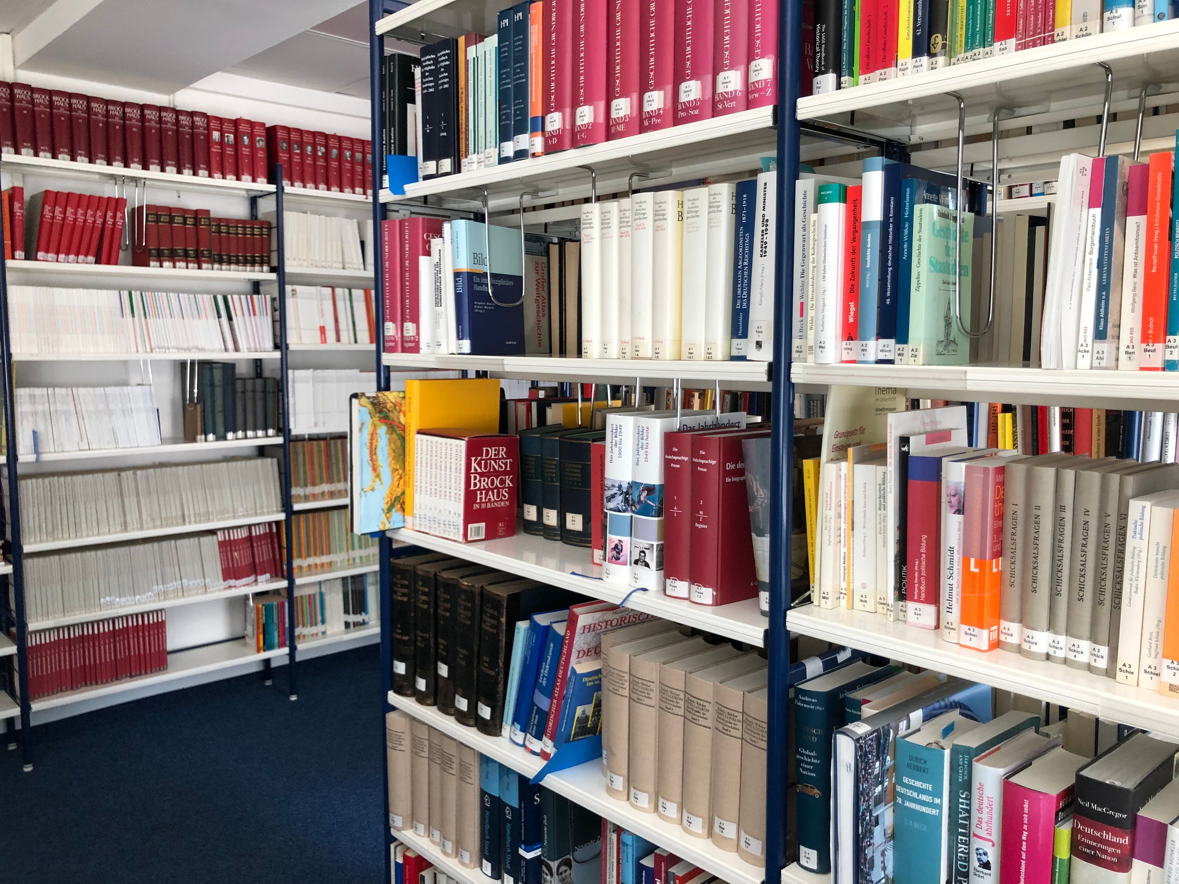 Innenansicht einer Bibliothek mit hohen, weißen Metallregalen voller Sachbücher und Lexika auf blauem Teppichboden.