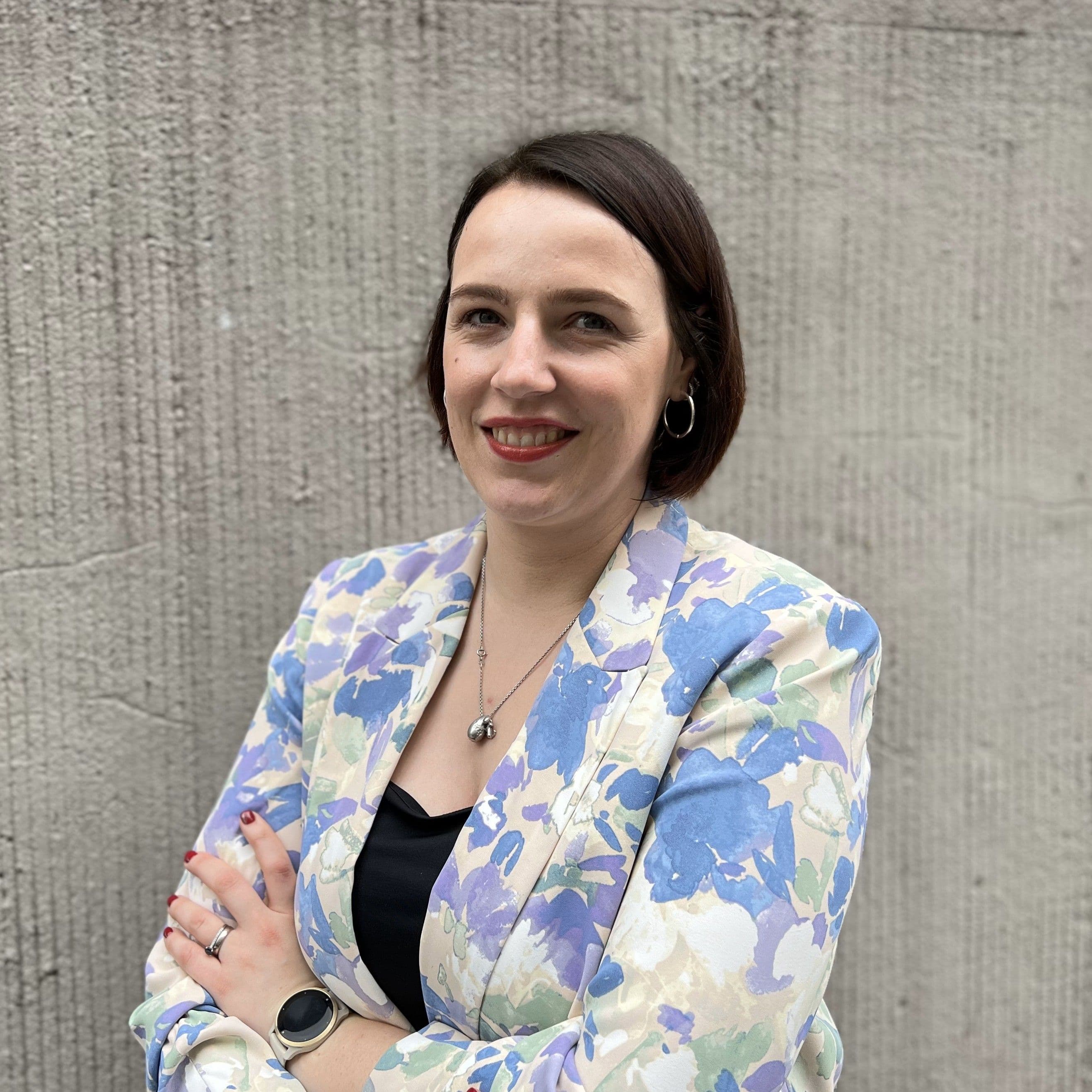 Porträt von Lisa Marie Freitag mit dunklem, kinnlangem Haar vor einer grauen Betonwand. Sie trägt einen weiß-blauen Blazer mit Blumenmuster, eine schwarze Bluse und eine Smartwatch.