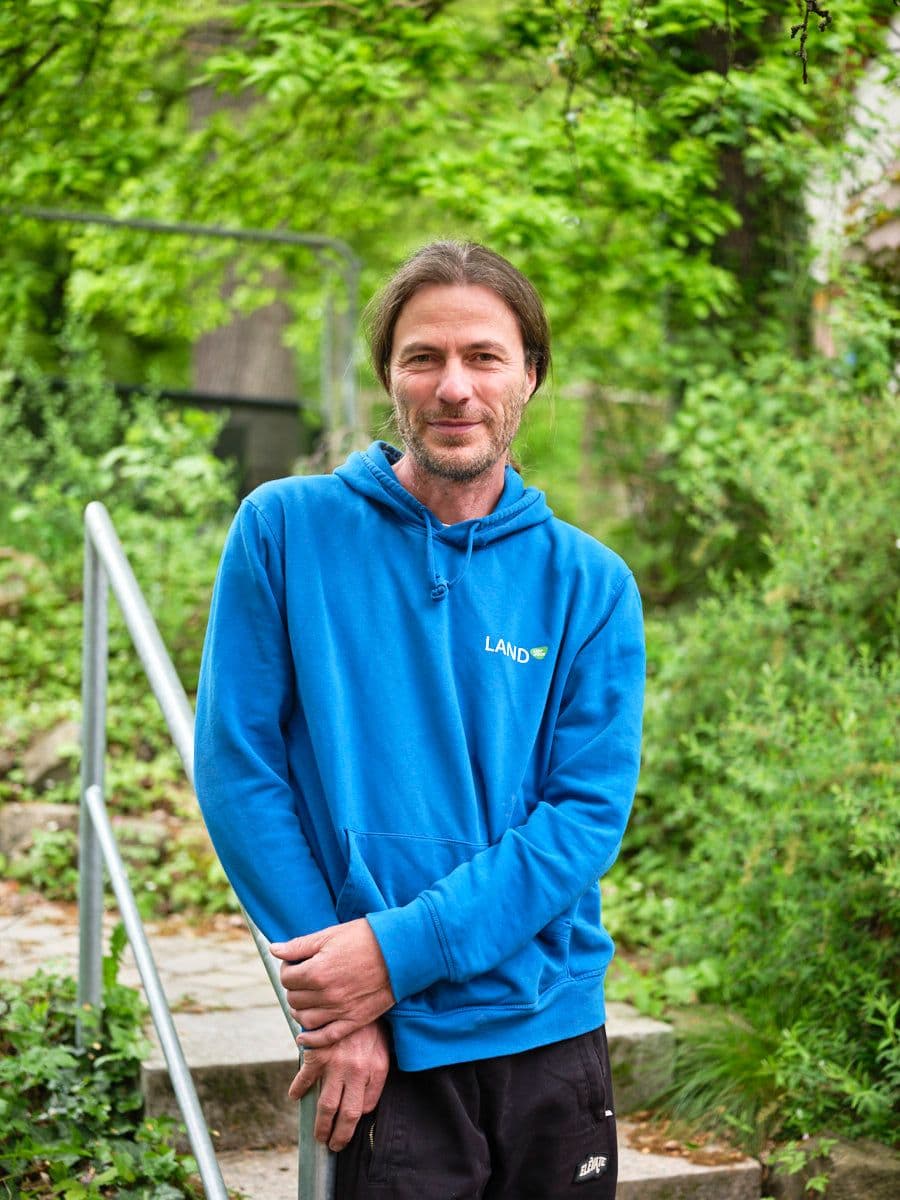 Porträtfoto eines Mannes mit zu einem Zopf gebundenen braunen Haaren und Dreitagebart, gekleidet in einen blauen Hoodie mit der Aufschrift „Land". Er hält einen angebissenen Apfel in der Hand und lächelt leicht in die Kamera. Der Hintergrund zeigt einen grünen Gartenweg mit Treppe und Geländer im Freien.