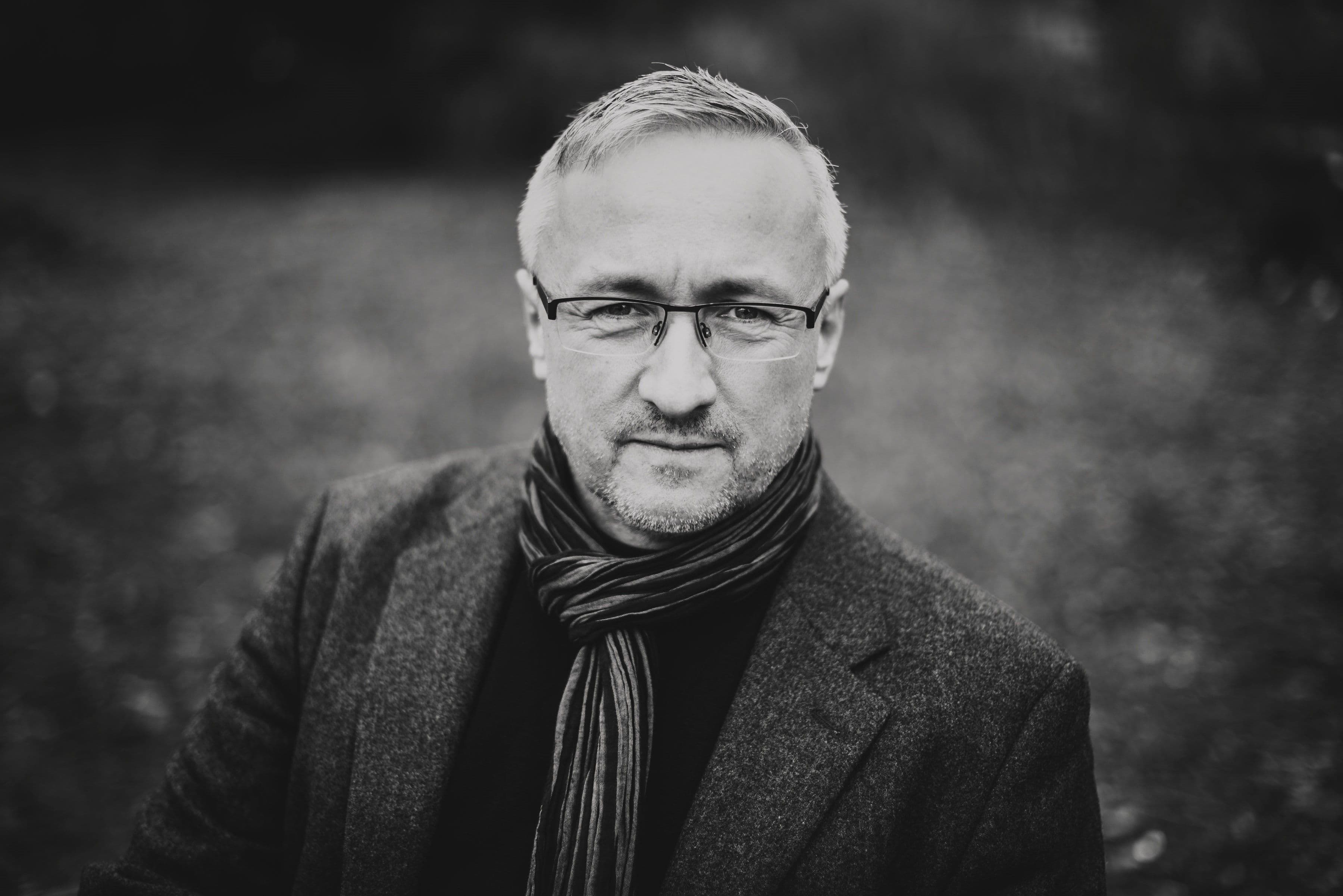 Eine Schwarz-Weiß-Nahaufnahme von Tobias Arand mit kurzen hellen Haaren und Brille. Er blickt direkt in die Kamera und trägt ein dunkles Sakko sowie einen gestreiften Schal. Der Hintergrund ist unscharf und zeigt eine natürliche Umgebung im Freien.