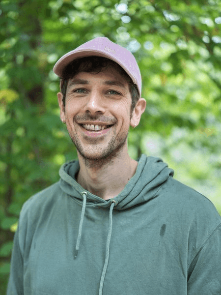 Porträtfoto eines Mannes mit einem rosafarbenen Cap und leichtem Bartansatz, gekleidet in einen olivgrünen Hoodie. Er lächelt breit in die Kamera. Der Hintergrund zeigt unscharfes grünes Laub im Freien.