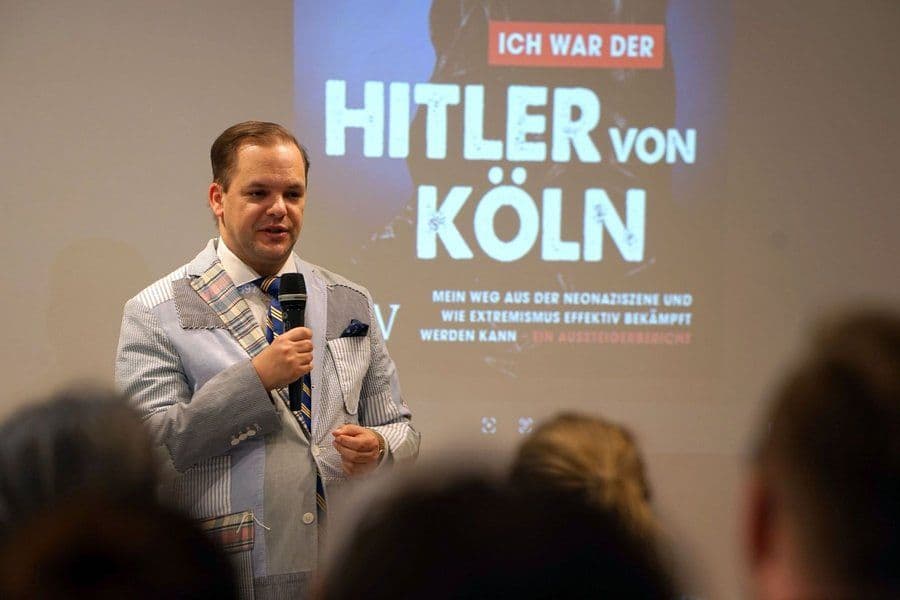 Mann mit Mikrofon spricht vor einem Publikum, im Hintergrund eine Präsentation mit dem Buchtitel "Ich war der Hitler von Köln"