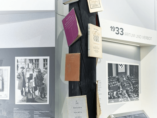 Museumsausstellung zum Thema „1933 – Irrtum und Verbot". Im Mittelpunkt steht eine schwarze Stele, an der mehrere historische Bücher befestigt sind, darunter verbotene oder verbrannte Werke der NS-Zeit. Links sind historische Schwarz-Weiß-Fotografien zu sehen, rechts ein Foto einer Parlamentssitzung. Die Ausstellungsgestaltung verweist auf die Bücherverbrennung und den Beginn der nationalsozialistischen Diktatur.