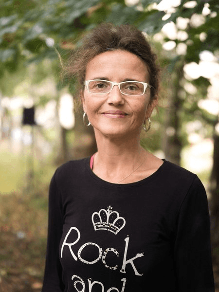 Porträtfoto einer Frau mit hochgesteckten dunklen Haaren und einer weißen Brille, gekleidet in ein schwarzes Langarmshirt mit dem Aufdruck „Rock and…". Sie lächelt leicht in die Kamera. Der Hintergrund zeigt unscharfe Bäume im Freien.
