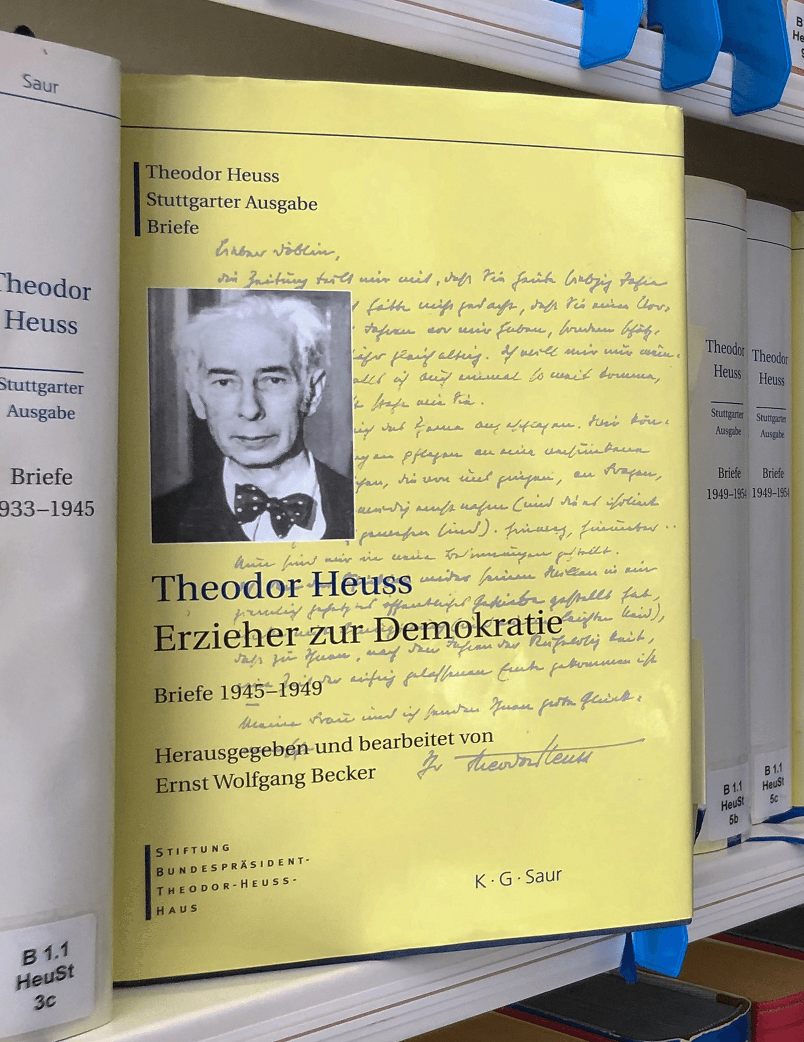 Ein Buchcover mit dem Titel „Theodor Heuss: Erzieher zur Demokratie“ steht im Vordergrund eines Bibliotheksregals, flankiert von weiteren Bänden der Stuttgarter Ausgabe.