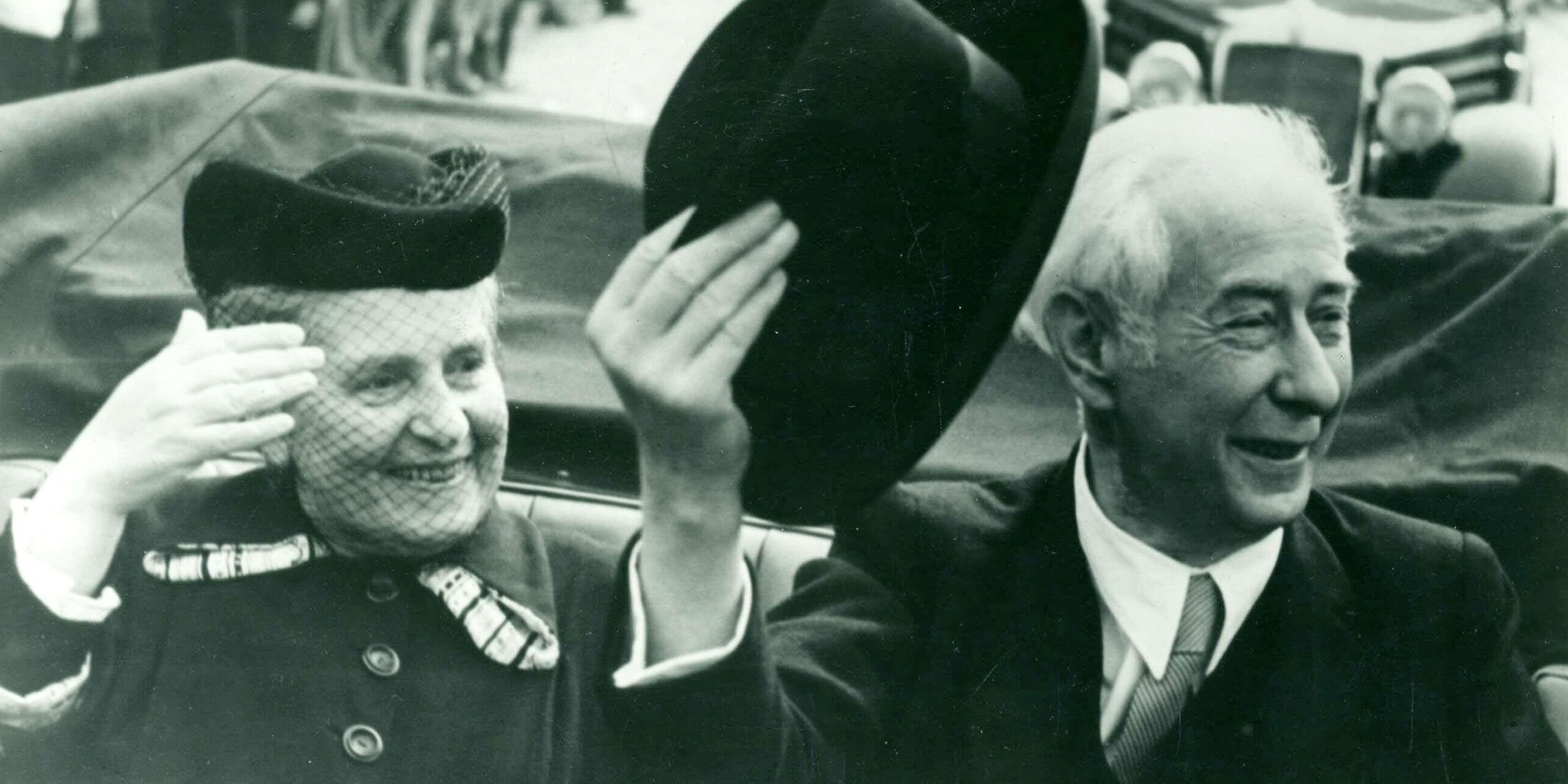 Theodor Heuss sitzt in einem offenen Auto, hebt einen Zylinder von seinem Kopf ab und grüßt. Neben ihm sitzt seine Frau Elly Heuss-Knapp.