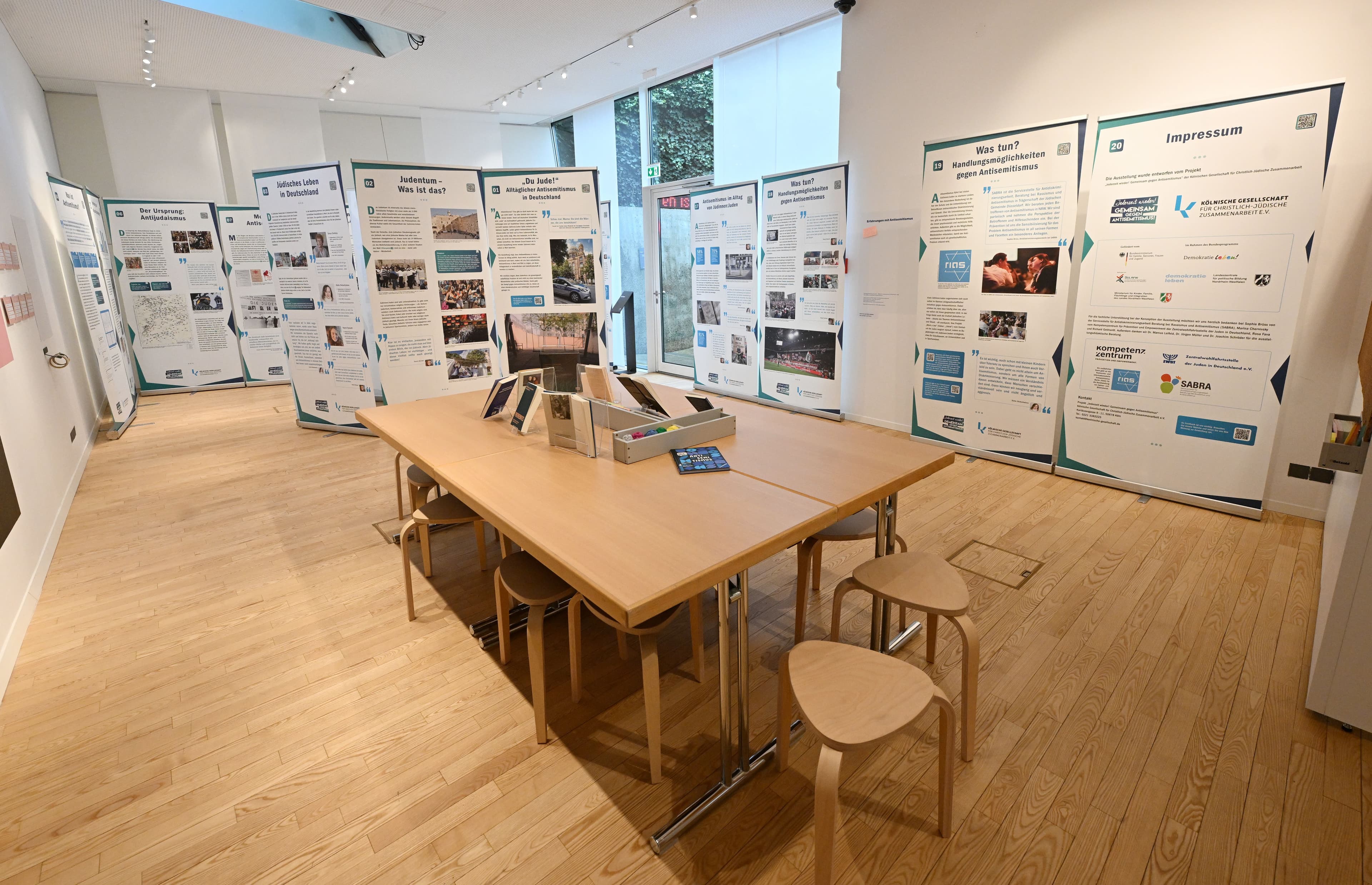 Ein heller Ausstellungsraum mit einem großen Holztisch in der Mitte, umgeben von zahlreichen Informationstafeln zum Thema Judentum und Antisemitismus.