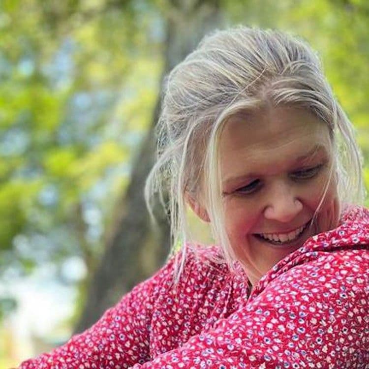 Porträtfoto einer Frau mit langen blonden Haaren, gekleidet in eine rote Bluse mit Blumenmuster. Sie lacht und blickt leicht nach unten. Der Hintergrund zeigt unscharfe Bäume im Freien.