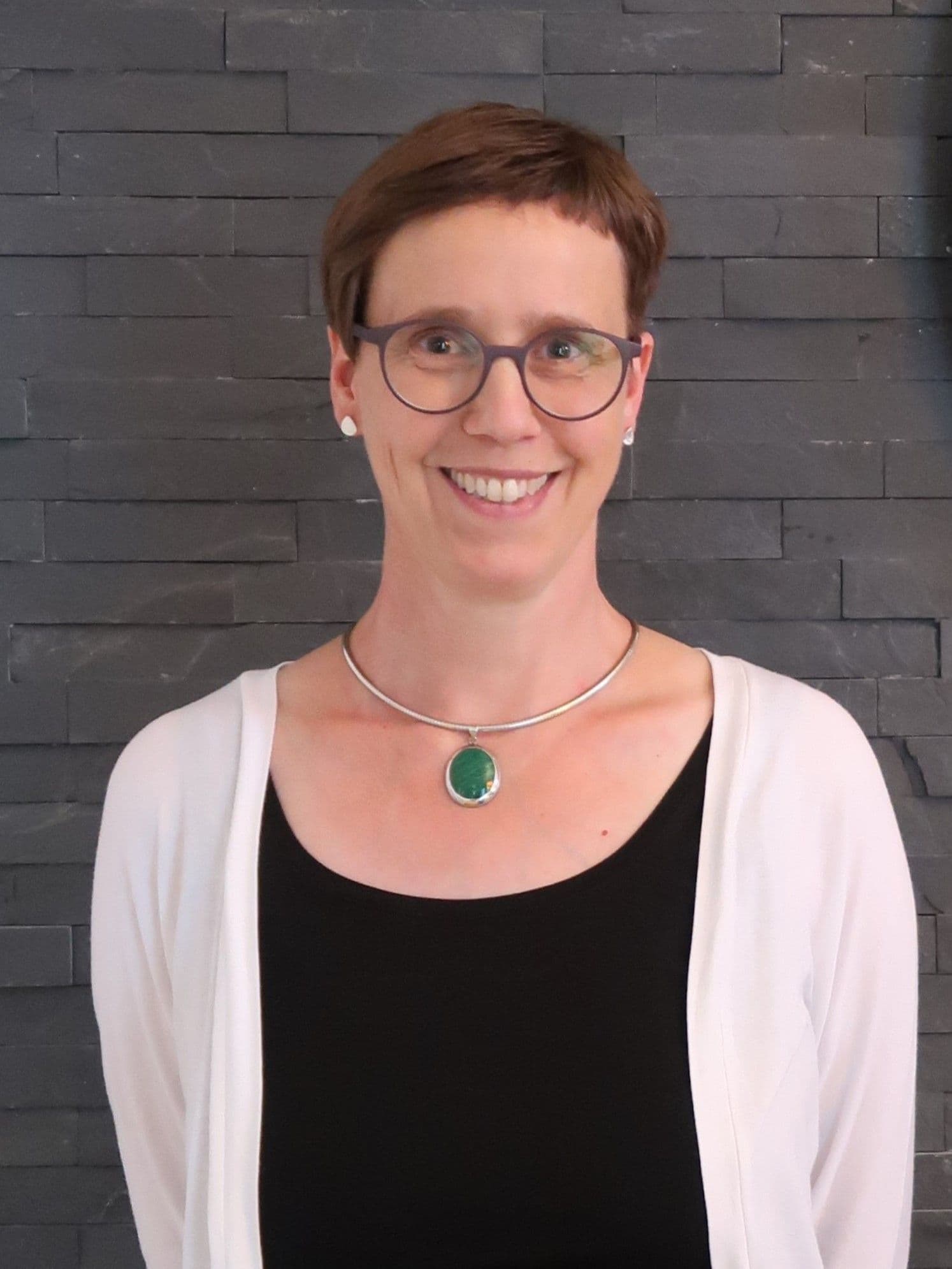 Porträt von Julia Schulze Wessel mit kurzen braunen Haaren und runder Brille vor einer dunklen Schieferwand. Sie trägt eine weiße Strickjacke über einem schwarzen Oberteil und eine Kette mit einem großen grünen Anhänger.
